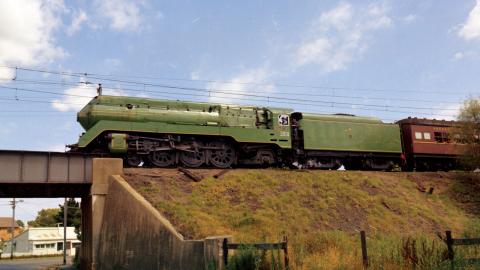 A 3801 steam locomotive pulling a passenger train on a raised railway track above a grass embankment. The train is about to cross a railway bridge