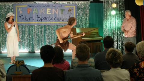 On stage at a school parents' talent show, a woman in a frilly hat and old-fashioned nightgown holds the microphone while a man in a loincloth holds a spear and lunges towards the compere standing in the wings.