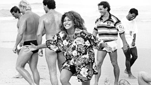 Tina Turner laughing on a beach, surrounded by male rugby league players in swimming costumes.