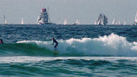 Two surfers in the ocean catching waves, in the background are tall ships and sailing boats.