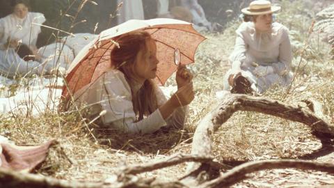 Miranda rests on the grass under a parasol and looks through a magnifying glass in a scene from the film Picnic at Hanging Rock