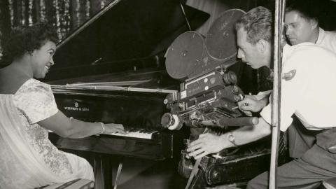 Winifred Atwell playing the piano and smiling while two men film her with a 35mm film camera next to the piano