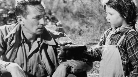 Peter Finch sitting on the ground holding his hat talking to a child