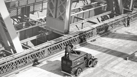 Fox Movietone Productions van with a man standing on top operating a camera. The van is on the deck of the newly-constructed Sydney Harbour Bridge.