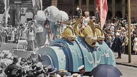 A partly colourised photo of a nautical-themed float in a 1930s street parade. On the float a woman dressed in white sits on a large-scale model of a sea creature.