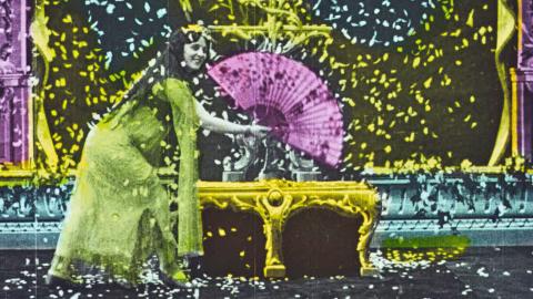 Still from a colourised film showing a woman bent down near a table holding a large fan.
