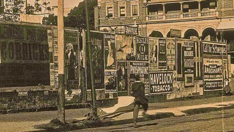 A scene from the early silent film Bashful Mr Brown shows a man walking down a street next to a fence lined with posters and advertisements.