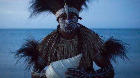 An indigenous man in traditional feathered costume holding a large shell with the ocean in the background.