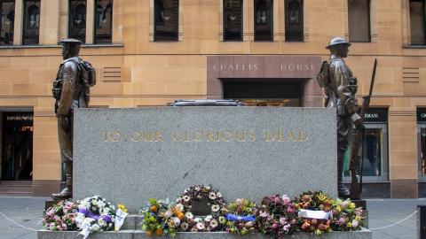 An Anzac Day memorial in Sydney inscribed 'To Our Glorious Dead' and flanked by statues of two soldiers. Wreaths of flowers surround the memorial