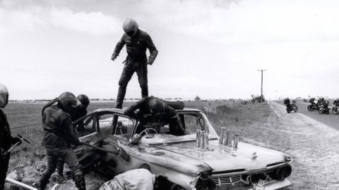 Members of a motorcycle gang smash up a car by the side of a highway