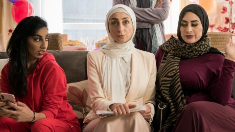 Three women sitting on a couch. One is looking sideways with an expression of annoyance at the other two women who are both looking straight ahead.