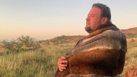 A man standing in profile pictured from the waist up wrapped in a traditional animal skin blanket.