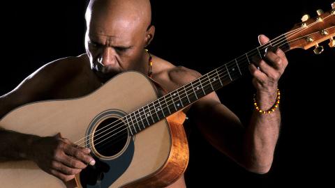 An Aboriginal man holding an acoustic guitar with his head bowed.