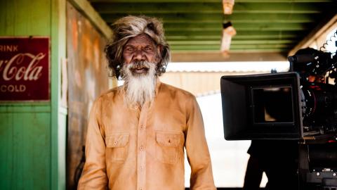David Gulpilil on the set of Satellite Boy with a film camera beside him. He is looking directly at the camera.