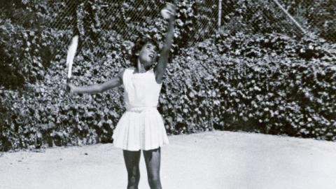 A young Aboriginal woman in a white tennis dress serving a tennis ball.