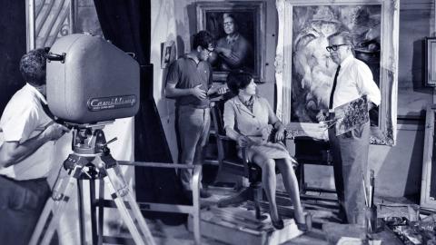 Blue-tinted black and white still of a man is operating a large film camera. He is filming artists Margaret Olley and William Dobell. There are two portraits painted by Dobell in the background as well as another member of the film crew.