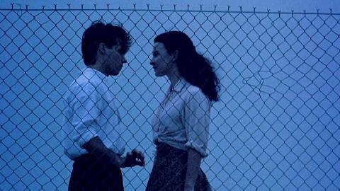 Paul Mercurio and Tara Morice in scene from the film Strictly Ballroom. They are facing each other in front of a wire fence. The time of day seems to be early evening. 