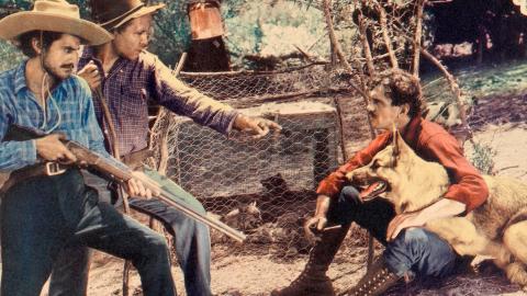 Two men hold rifles and stand over a man sitting on the ground with a dog
