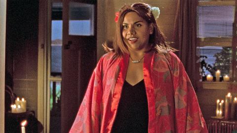 A young Aboriginal woman wearing a red kimono and flower in her hair stands in the middle of a candlelit room facing camera with a half-smile on her face.