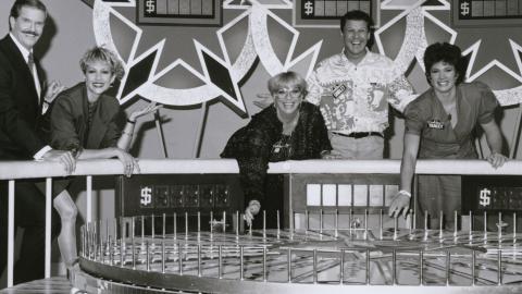 Game show hosts and contestants in a TV studio standing behind a big spinning wheel.