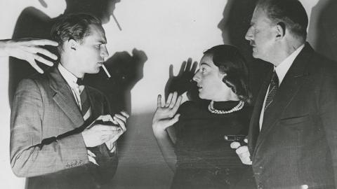 Two men and a woman. One man lights a cigarette while a mysterious hand reaches for him. The woman looks terrified. The other man holds a gun.