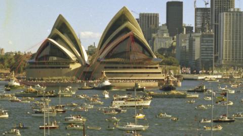 The Sydney Opera House is festooned with red ribbons. There are many boats on the water in front of it.