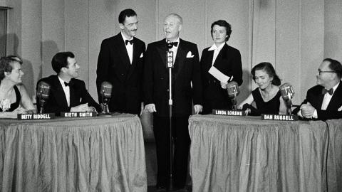 A studio with 3 people standing in the centre in front of a radio microphone and a table on each side with two panellists seated at each table.