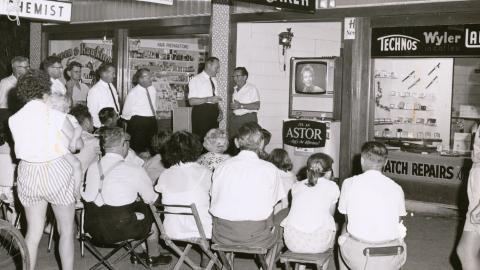 A crowd of people (some seated and some standing). They are watching a television set through a shop window.