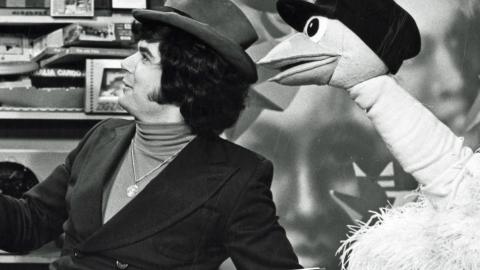 A man wearing a hat sitting next to an ostrich puppet who is also wearing a hat. They are both turned to their right.