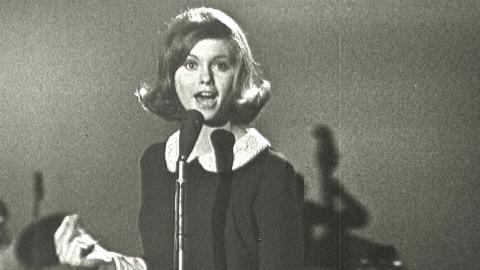 Olivia Newton-John at age 16 in a TV studio standing behind a microphone singing.