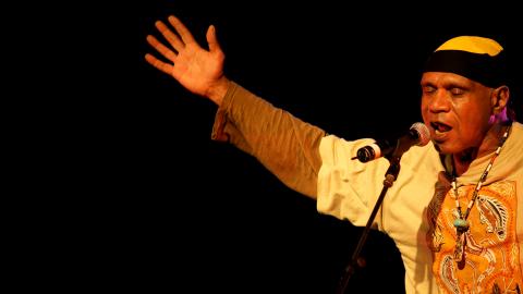 Archie Roach stands at a microphone singing. He is holding his arms outstretched.
