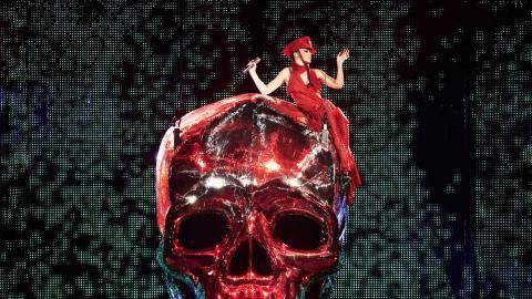 Kylie Minogue on red skull Kylie Minogue, dressed entirely in red, is sitting on top of a giant red shiny skull prop at one of her live shows.