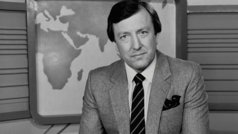 Journalist Jim Waley is wearing a suit and tie in this black and white photo. He is sitting in a studio with a backdrop showing a map of the world.