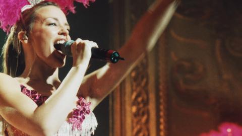 Kylie Minogue on stage in a pink showgirl costume singing into a microphone.