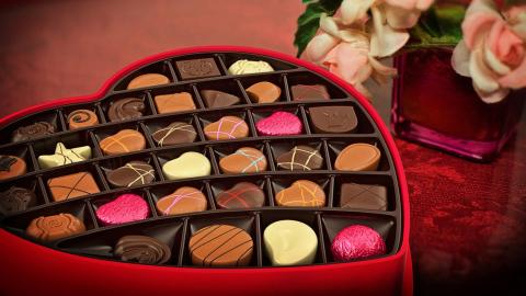 A heart shaped box of assorted chocolates with a small vase of pink roses in the background