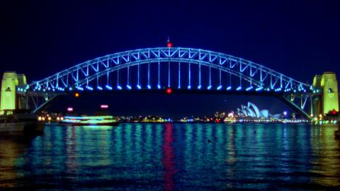 A nightime photo of the Sydney Harbour Bridge showing it all lit up. The bridge arch is blue with red lights at the top and bottom centre. The pylons are lit yellow. The water shows the reflections of the passing ferry and the Sydney Opera House.