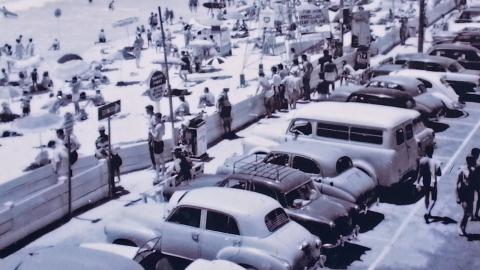 A busy Australian beachfront showing people on the sand and cars behind the sand in a parking lot, circa 1960s.