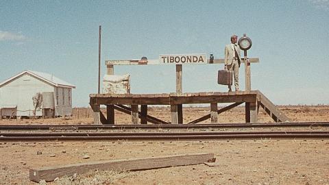A man carrying a suitcase and wearing a light brown suit waits on a small outback railway platform