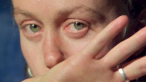We Aim to Please A woman looks at the camera as she wipes her face with her hand.