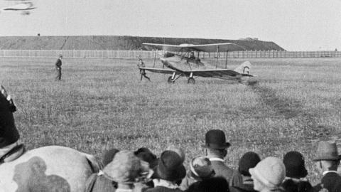 An early 20th century biplane lands in a field, watched by a crowd of spectators