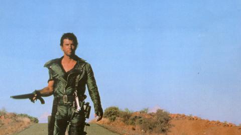 Mad Max walking resolutely down the middle of a desert highway, gun in hand