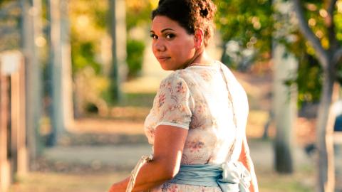 A woman in a floral dress pictured from the waist up, walking away but with her head turned to look back over her shoulder.