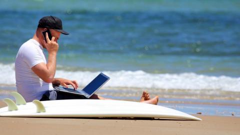 A man working on the beach with a mobile phone and laptop and a surfboard on the sand next to him