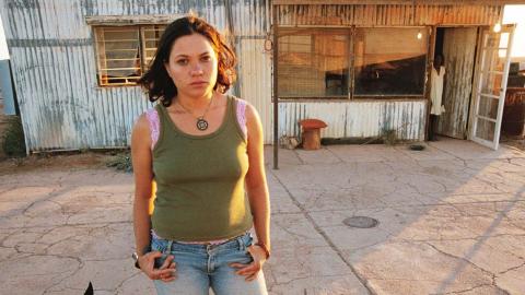 A young woman stands outside a corrugated iron building; an older Aboriginal woman stands in the doorway behind her