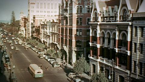 A street scene from Perth circa 1954. A street scene from Perth circa 1954. The street is lined with 3 and 4 story buildings and there are many cars parked on the side of the road, there are trees along the footpaths and a few pedestrians can be seen crossing the street.
