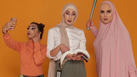 Three young women - two wearing the hijab - stand next to each other. One is taking a mobile phone selfie, another in corporate dress holds a law book, while the third wields a baseball bat.