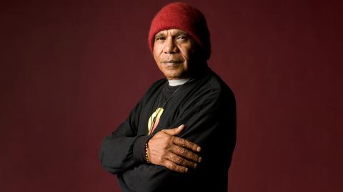 Archie Roach looks at the camera with his arms folded.