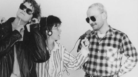 A woman wearing headphones aims a radio microphone on a cord at a man standing next to her. Another person leans against the woman, as if listening in on their recorded conversation.