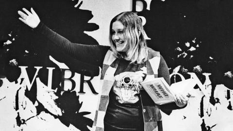 Journalist Lillian Roxon waving at a crowd. She is holding a book.