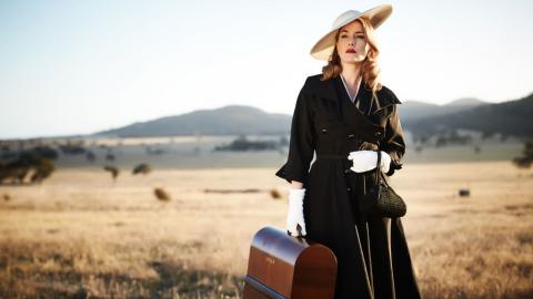 A woman in a black coat and hat holding a suitcase in the Australian outback